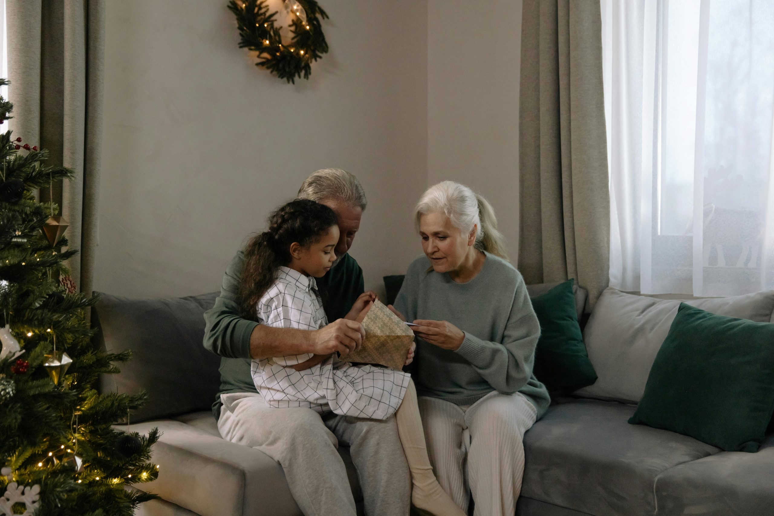 an elderly couple sits with their granddaughter on the couch, opening a christmas present by a christmas tree